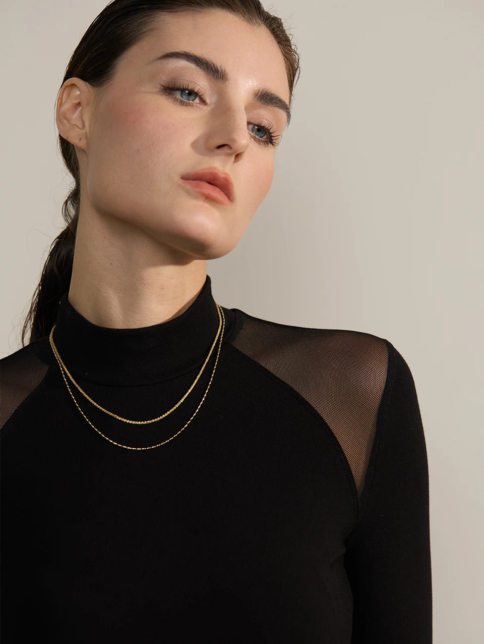 Woman wearing a black sheer top with a gold necklace against a neutral background