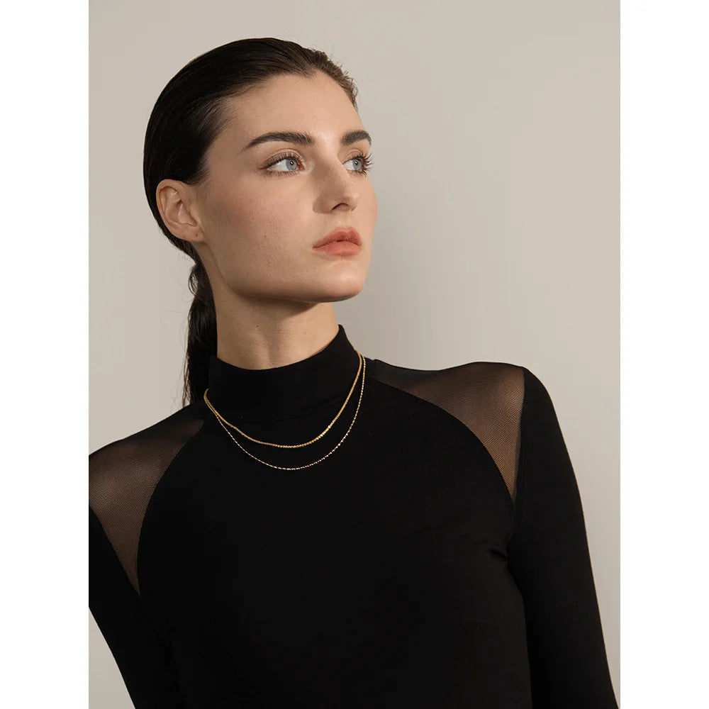 Woman wearing a black sheer top with gold jewelry against a neutral background