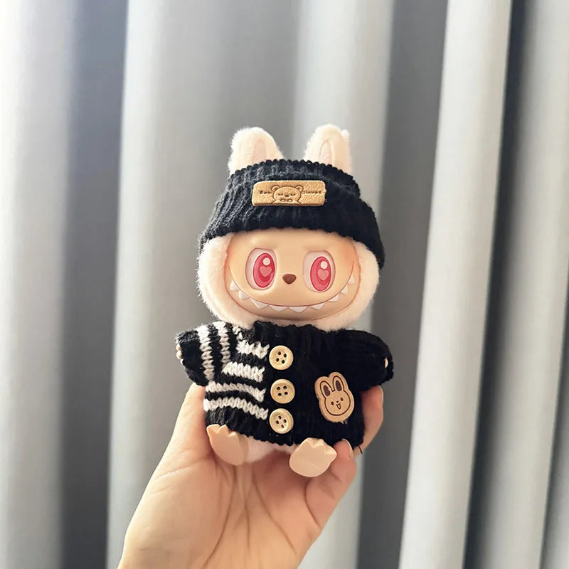 Small labubu  plush toy with a black knitted hat and sweater held in a hand against a neutral background