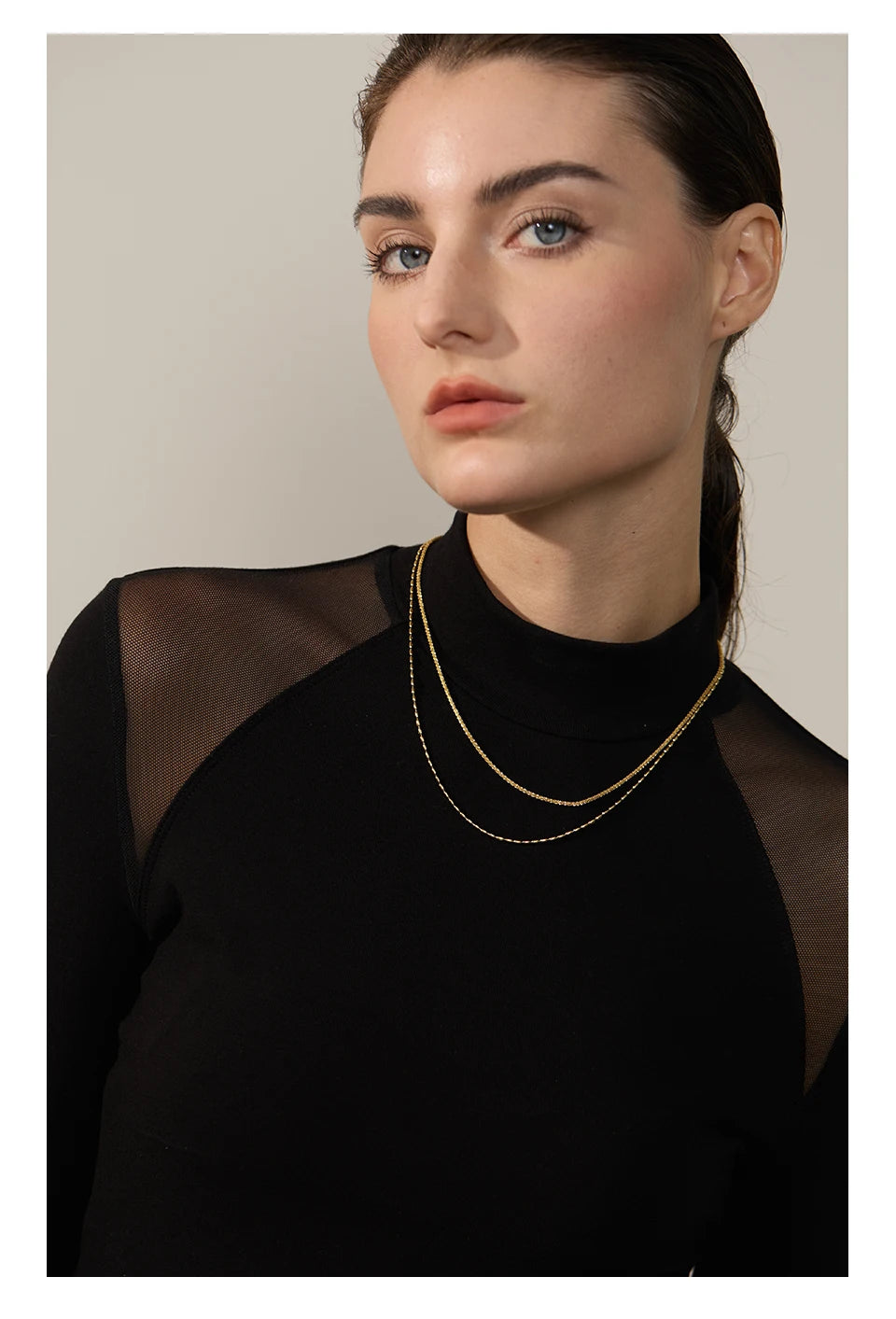 Woman wearing a black sheer top with a gold necklace against a beige background
