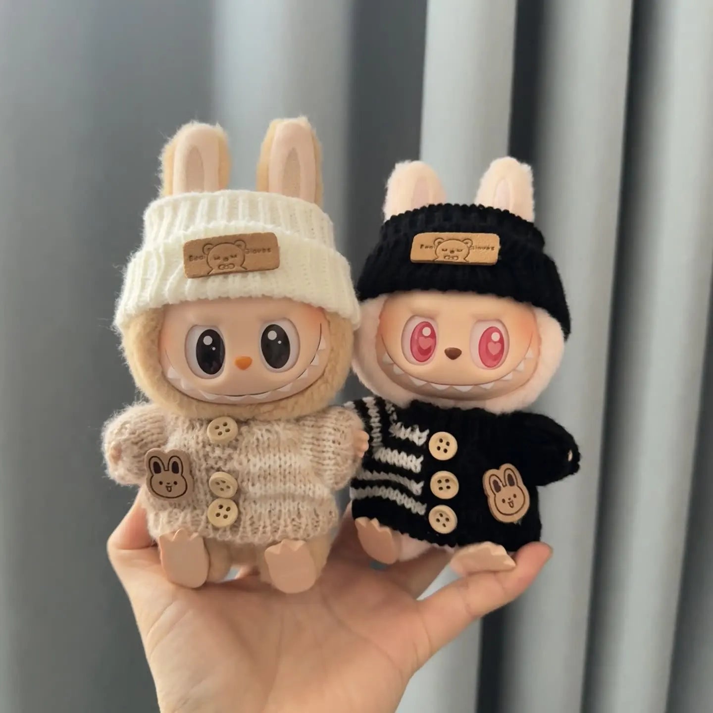Two labubu plush toys with hats and sweaters held in a hand against a neutral background